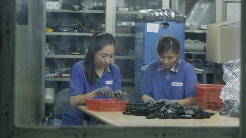 야간근무 스틸컷 4 공장 캐비넷에 온갖 물품들이 놓여져있다. 린과 연희는 근무복을 입고 손에 회색 장갑을 낀 채 가깝게 붙어 앉아 물품들을 조립하고 있다.