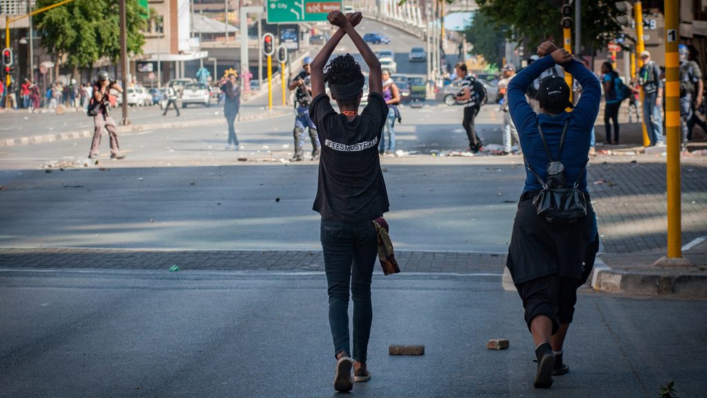 모든 것은 반드시 무너진다 스틸컷3. 시위에 참여한 두명의 시민이 머리 위로 손을 들고 손목을 엑스자로 만들어 걸어가고 있다. 촬영기사들은 이를 찍고 있으며 한 경찰이 멀리서 총을 들어 올리고 있다.