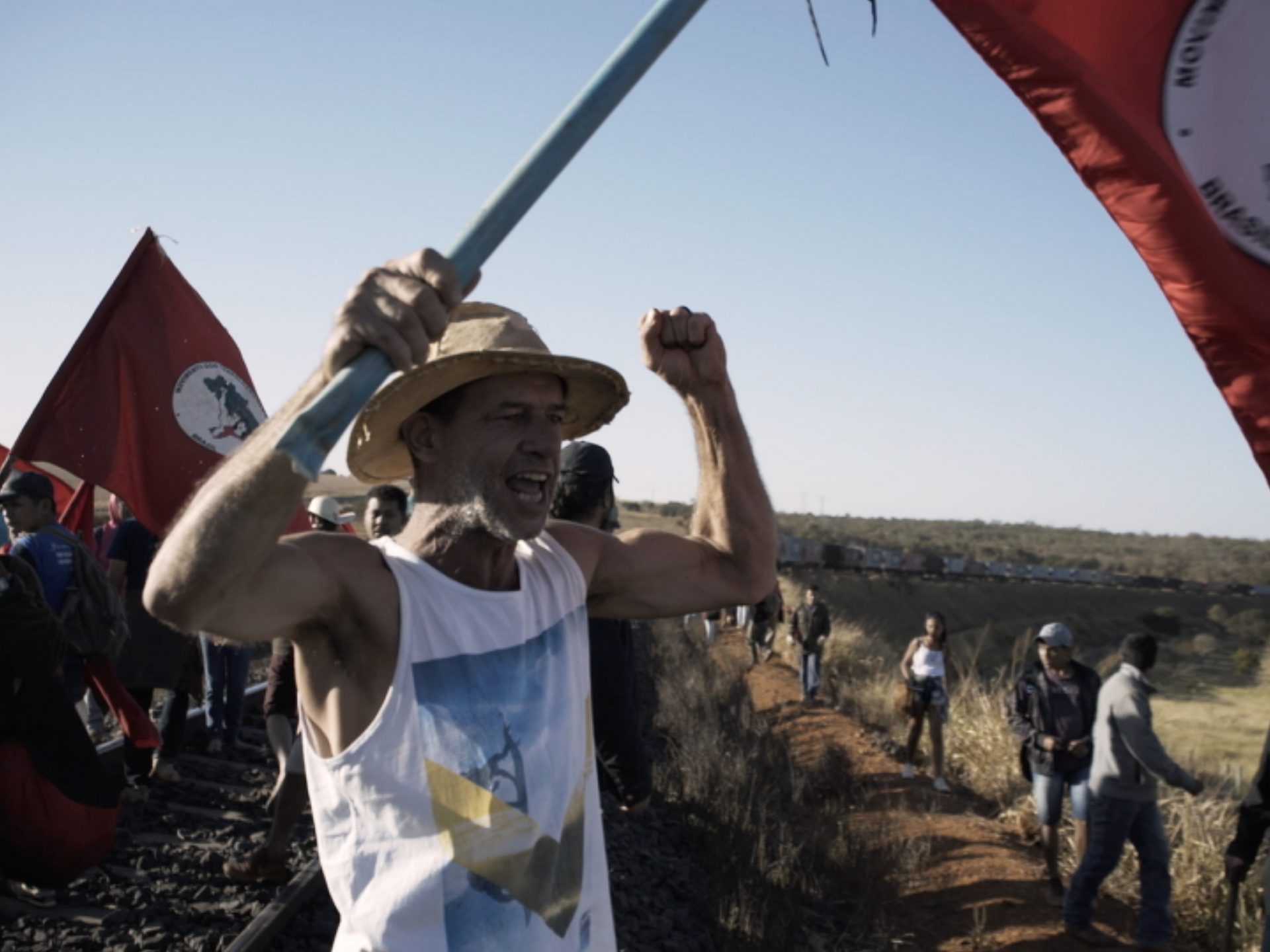 무토지 스틸컷4. 철도를 점령한 시위대가 깃발을 들고 서있다. 모자를 쓴 한 참여자는 한손에는 깃발을, 다른 한손으로는 주먹을 꽉 쥔채 구호를 외치고 있다.