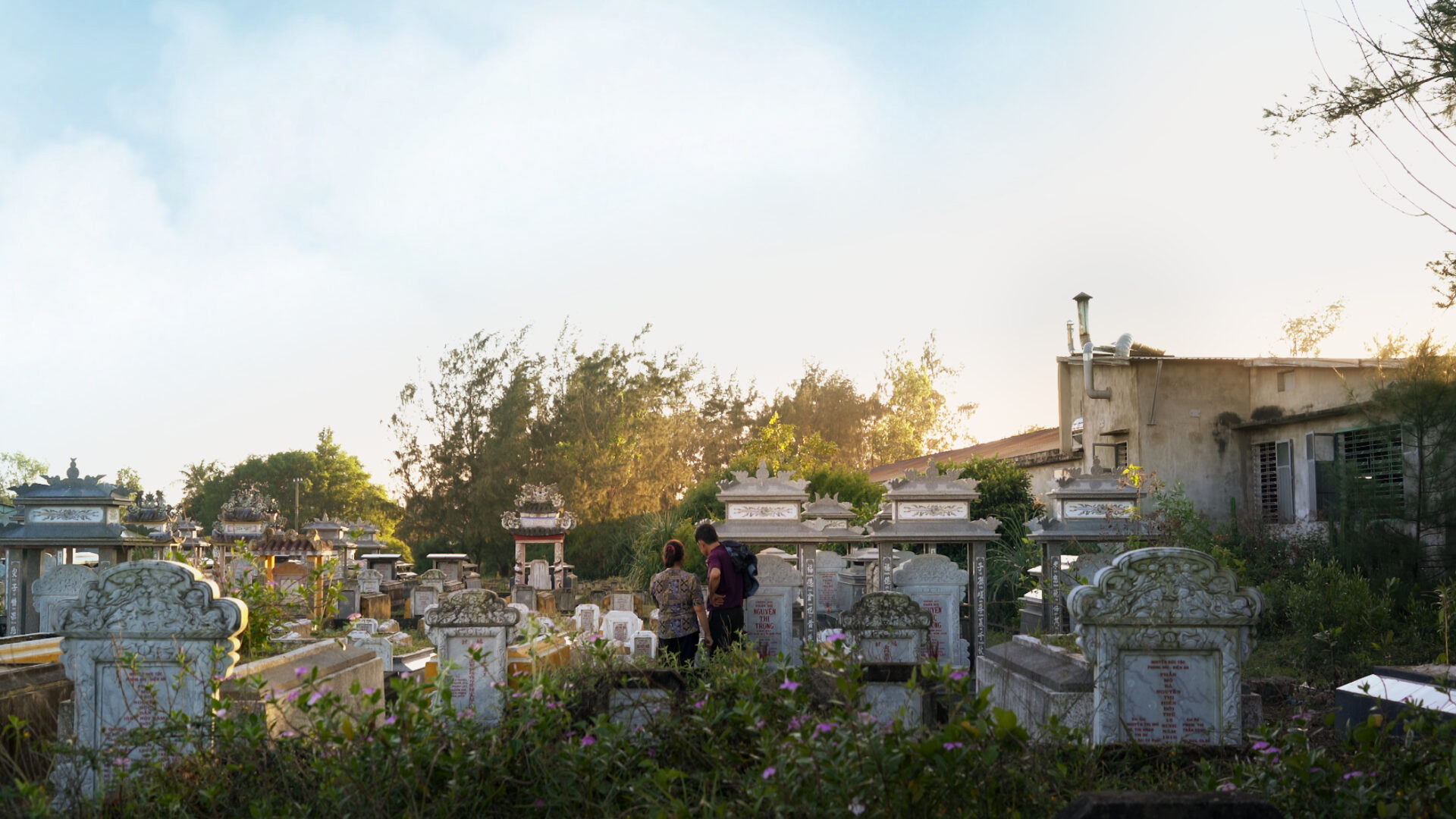 기억의 전쟁 스틸컷3. 햇살이 가득한 오후, 집단 묘지 가운데에 두 사람이 등을 보이고 서 있다. 뒤 쪽에는 나무들이 우거져 있다.