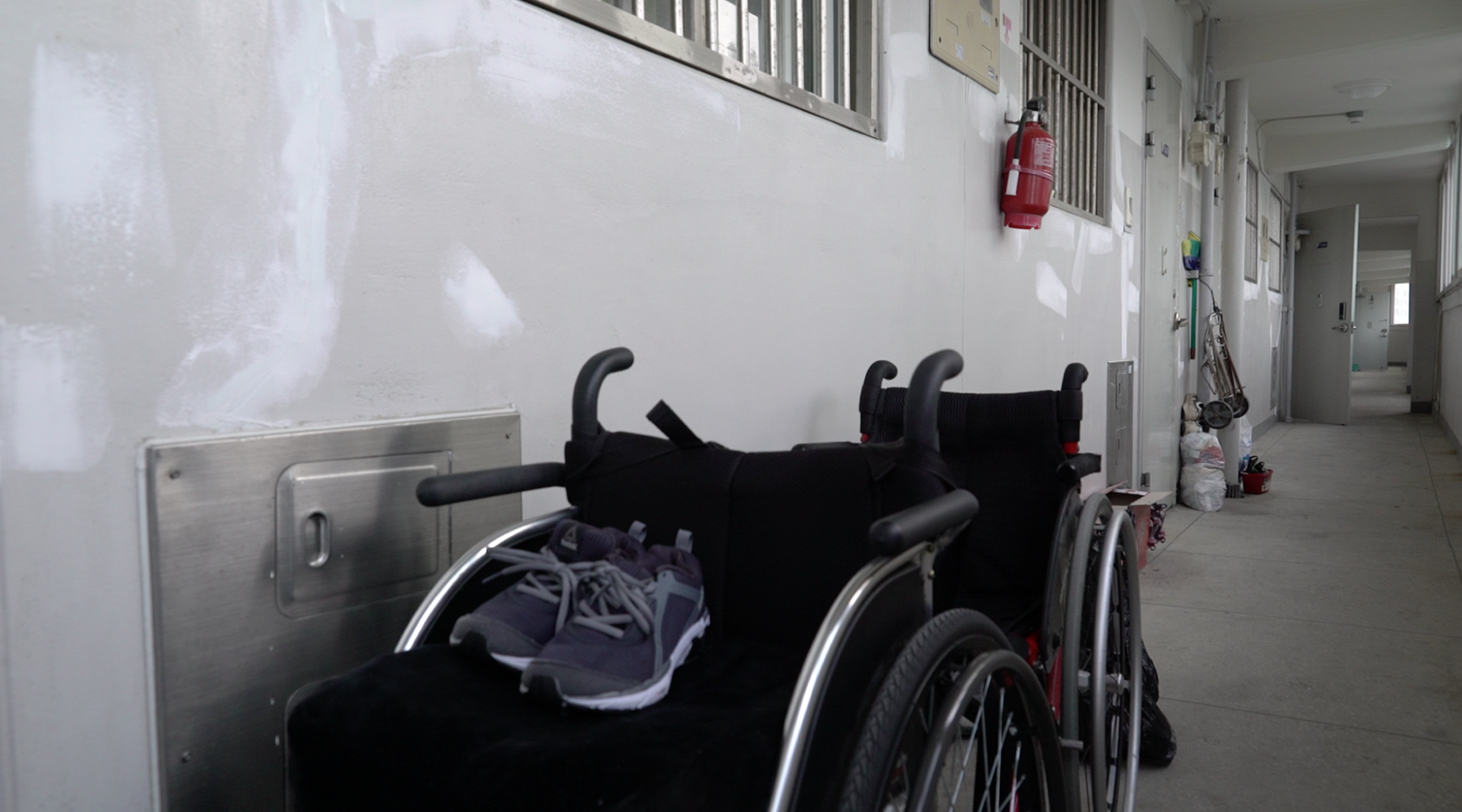 누구도 남겨두지 않는다 스틸컷2. 긴 아파트 복도 앞 쪽에 휠체어 두 개가 나란히 있다. 앞에 있는 휠체어 위에 남색 운동화 한켤레가 놓여 있다.