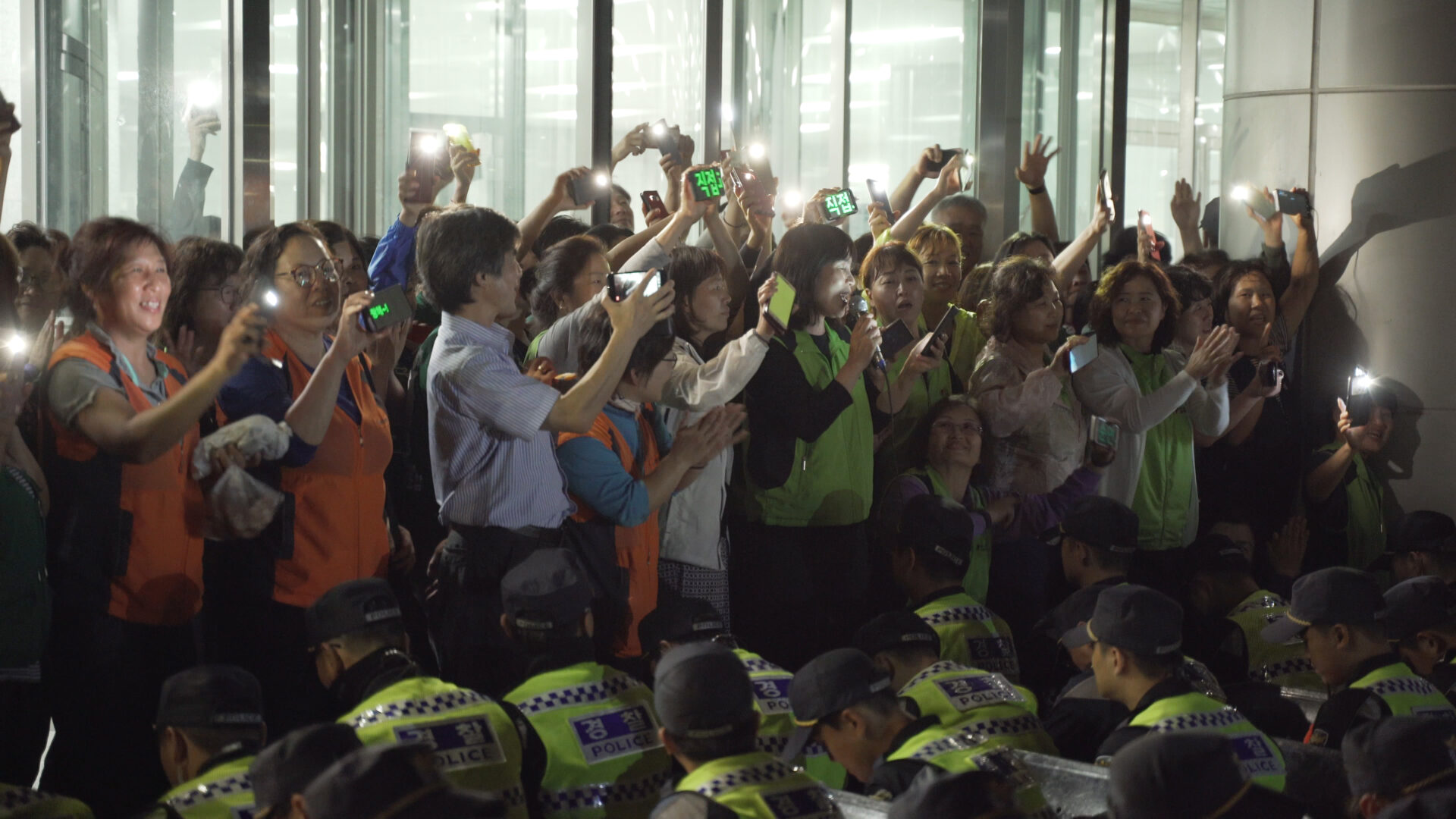 보라보라 스틸컷1.해가 진 저녁, 한국도로공사 김천본사 앞에서 노동자들이 핸드폰 불빛과 후레쉬를 킨채 손을 흔들며 웃고있다. 한 노동자는 마이크를 들고 웃으며 사회를 보고 있다. 이들의 앞에는 경찰대오가 줄을 맞춰 앉아있다.