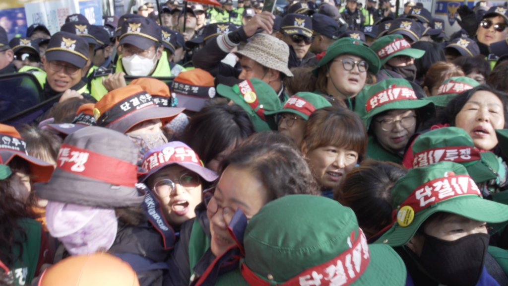 보라보라 스틸컷. 톨게이트 요금수납원들이 초록색, 주황색 모자 위에 ‘단결 투쟁’이라고 적힌 빨간 띠를 두른 상태로 경찰과 대치중이다. 발디딜 공간이 없을 정도로 빼곡하게 껴있는 노동자들은 얼굴을 찡그리고 있다.