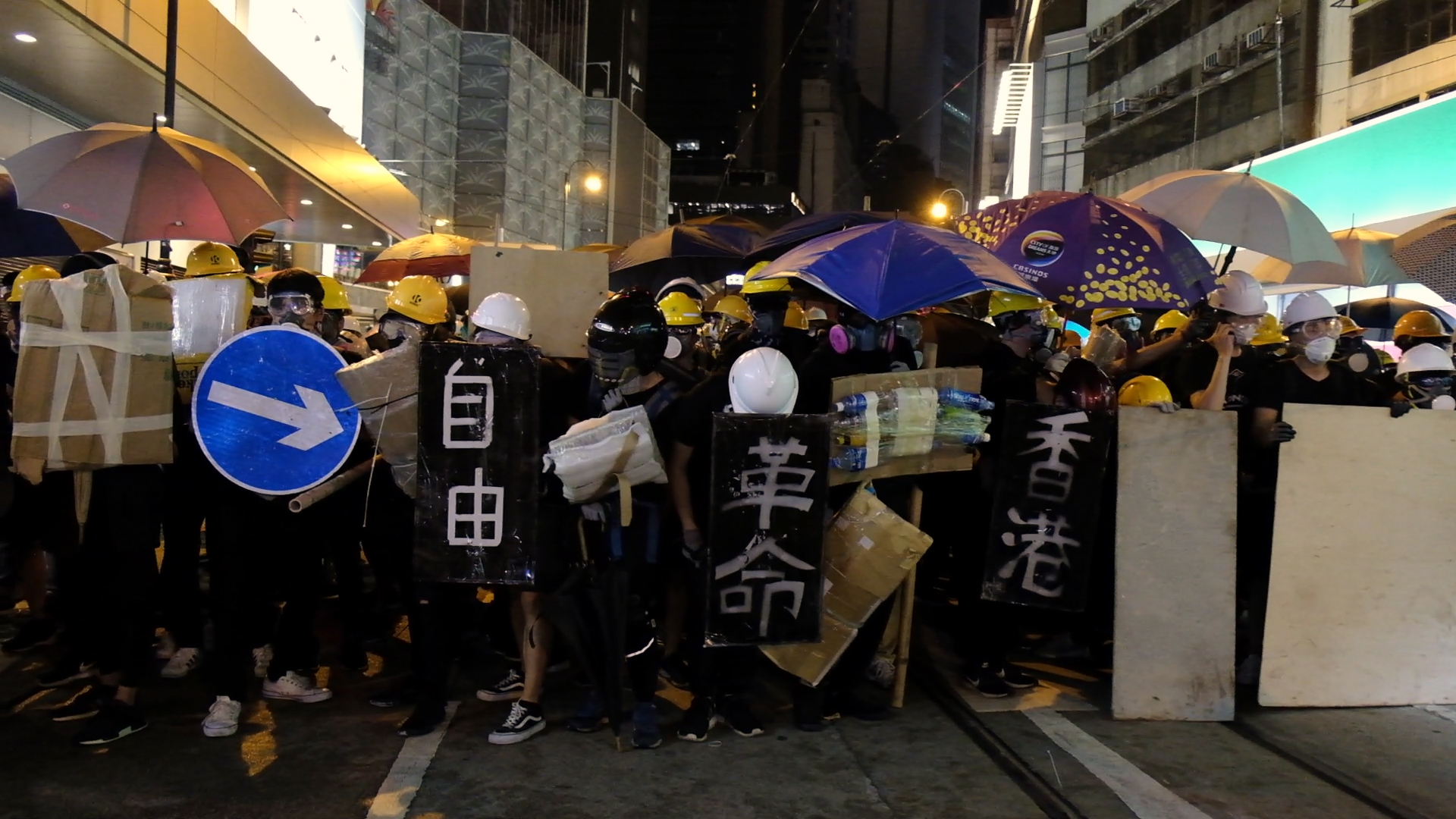 동지 스틸컷1. 밤,시위대가 검은 바탕에 흰색 글씨가 쓰여진 피켓을 든 채 정면을 향해 서 있다.