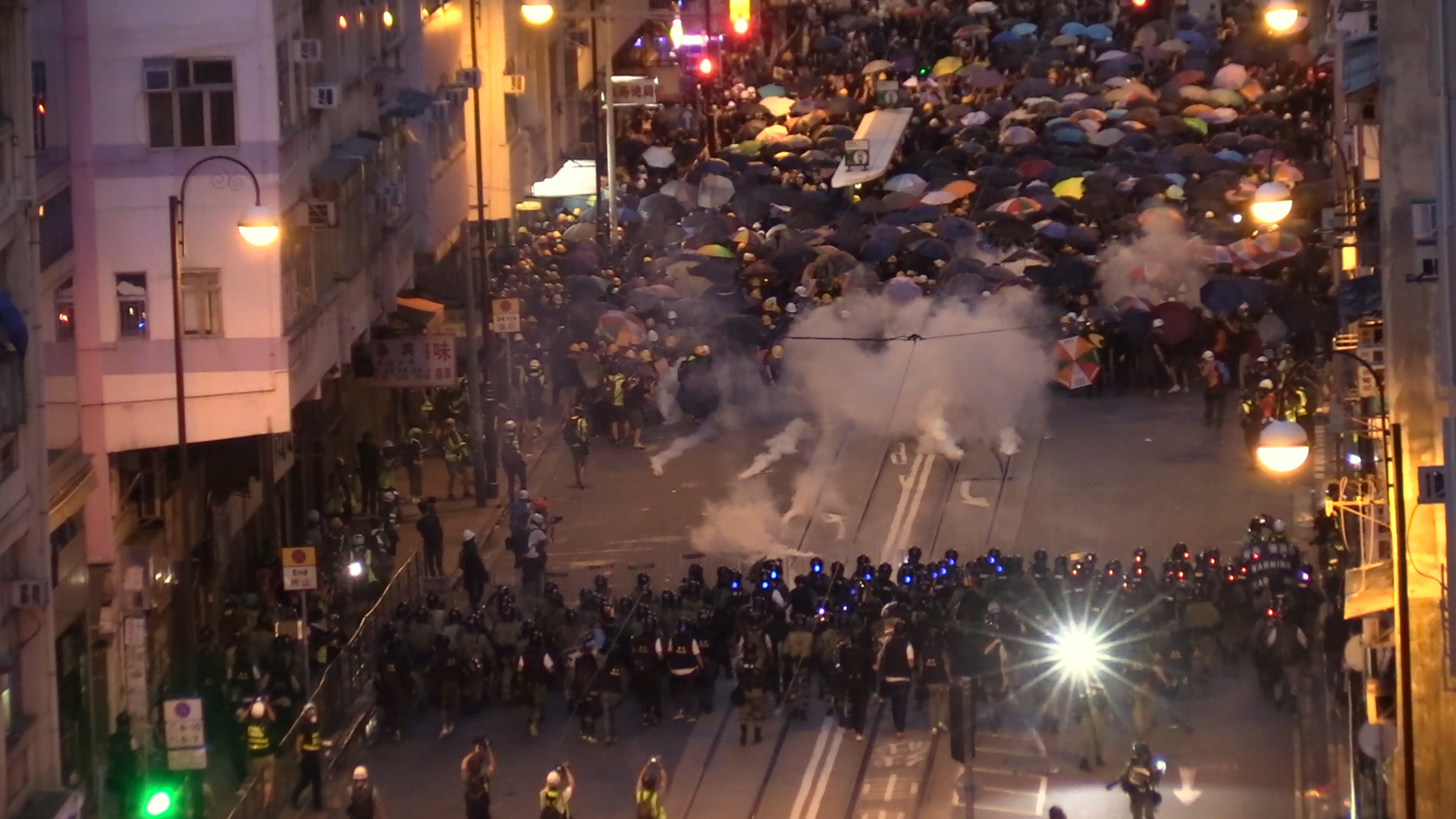 동지 스틸컷3. 밤 거리, 화면 위쪽의 우산을 펼쳐 든 시위대와 아래쪽의 경찰들이 대치하고 있다. 시위대와 경찰 사이의 빈 거리에서 연기가 피어 오른다.