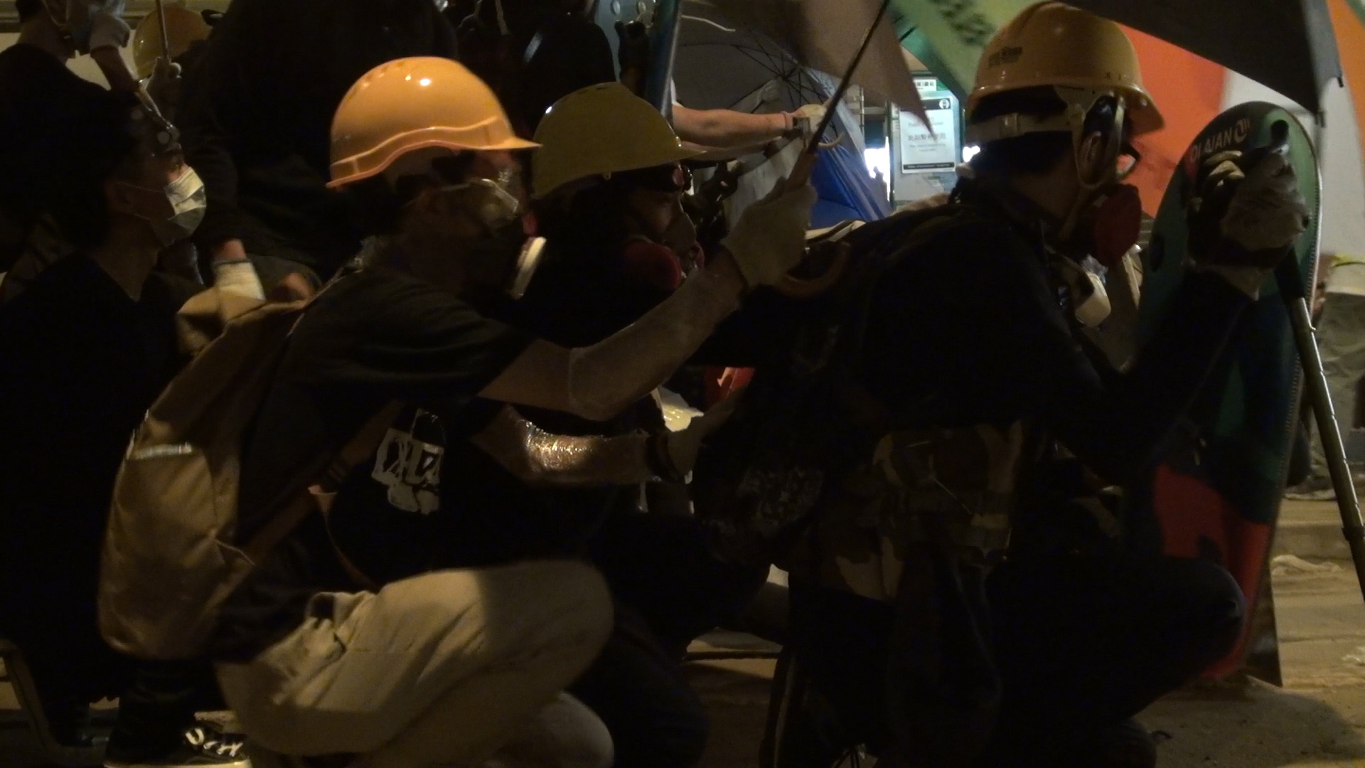 동지 스틸컷5. 헬멧을 쓴 집회 참가자들이 앉아 있다. 한 사람이 뒤에서 앞 사람을 우산으로 가려주고 있다.
