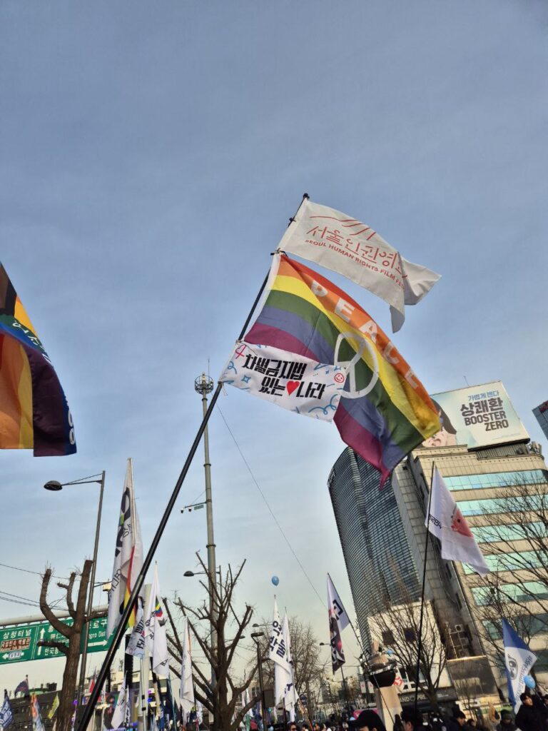 사진. 광화문 앞 집회에서 많은 깃발 가운데 서울인권영화제 깃발이 펄럭이고 있다. 서울인권영화제 깃발 아래는 무지개 깃발과 차별금지법 있는 나라 깃발이 같이 펄럭이고 있다.