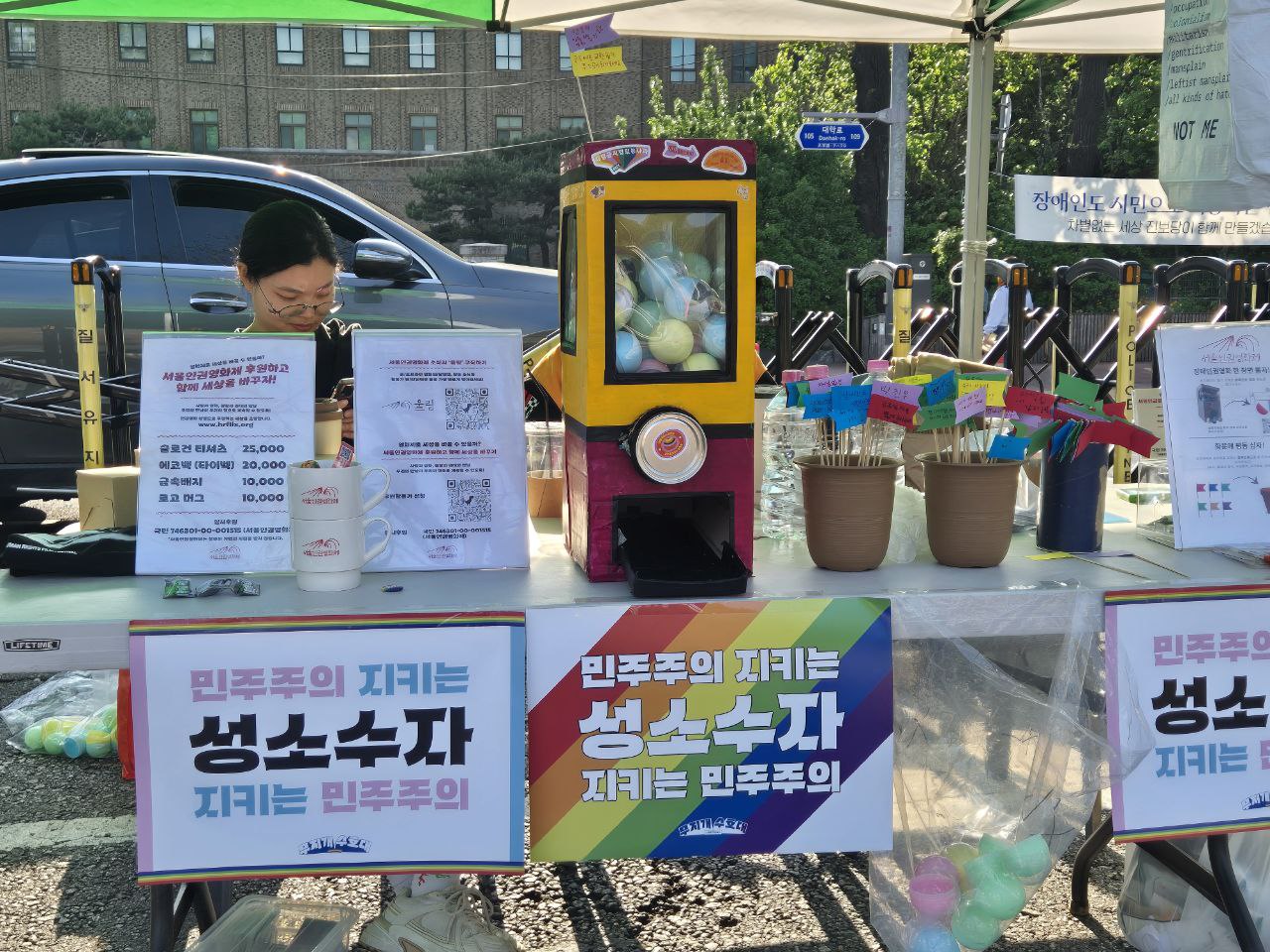 사진. 나기가 장애인차별철페의 날 행사 부스를 지키고 있다. 부스 테이블 위에는 안내판, 서울인권영화제 굿즈, 수제 뽑기 머신, 깃발이 꽂힌 화분 등이 놓여있다.