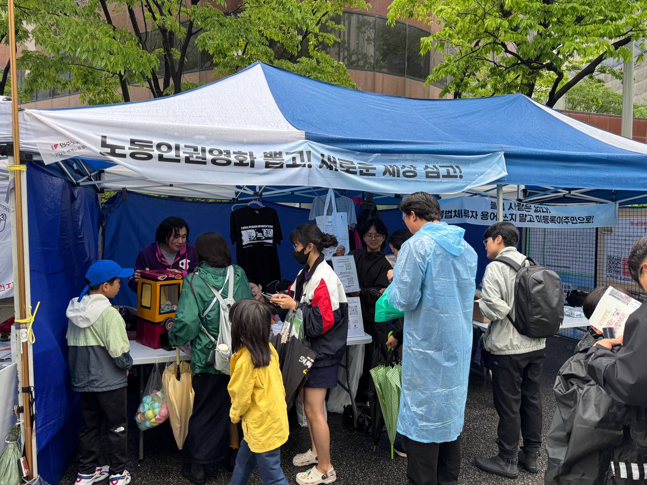 사진. 부스 천막에 ‘노동인권영화 뽑고! 새로운 세상 심고!’라고 쓰여있는 현수막이 붙어있다. 부스 안 왼쪽에서 활동가 소하가 네 명의 사람들에게 ‘서인영 가챠’ 방법을 설명하고 있다. 소하 옆에서 활동가 나기와 두부가 3명의 사람들에게 서울인권영화제 부스를 소개하고 있다. 사람들은 우비를 입고 있거나 우산을 들고 있다.