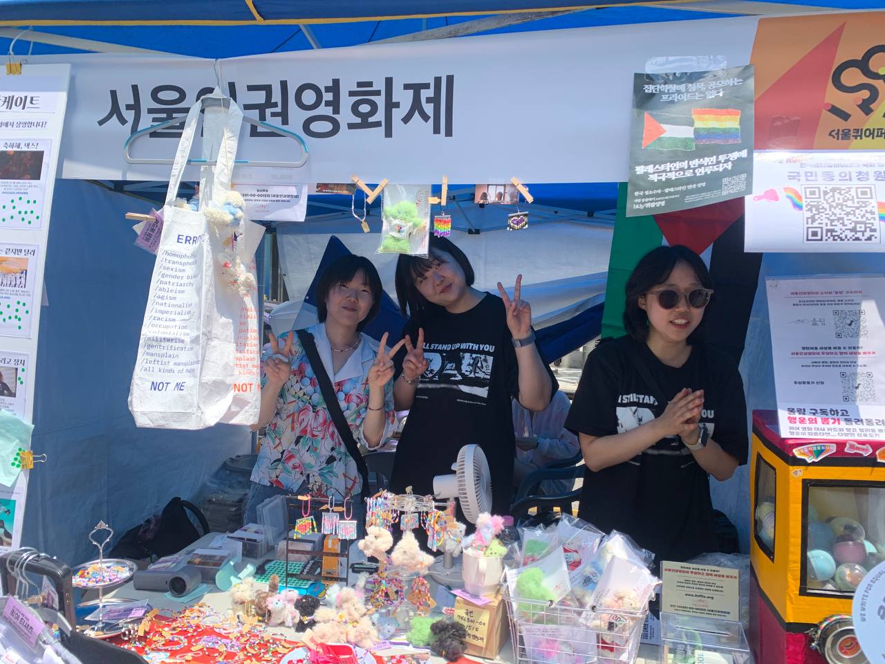 사진. 서울퀴어퍼레이드 부스에서 마주, 안나 고운이 포즈를 취하고 서 있다. 부스에는 각종 후원상품들이 진열되어있다.