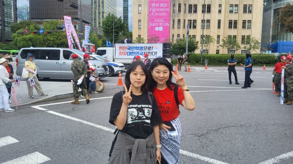 사진2. 자유통일당 집회에 견학(?) 간 고운과 민희(플랫폼C 활동가, 행진팀장)가 브이 포즈를 하고 서있다. 뒤편에 ‘천만국민혁명으로! 자유대한민국지킨다!’라고 적힌 현수막이 붙은 트럭이 있다.