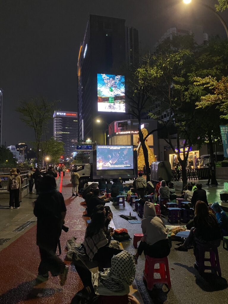 사진1. LED트럭 스크린에서 영화 “끝나지 않는 밤”이 상영 중이다. 참가자들은 바닥에 작은 플라스틱 의자와 깔개 등을 깔고 우산, 우비 등을 쓴 채로 영화를 보고 있다. 트럭 저 뒤편으로 주한이스라엘대사관이 있는 빌딩이 있다.
