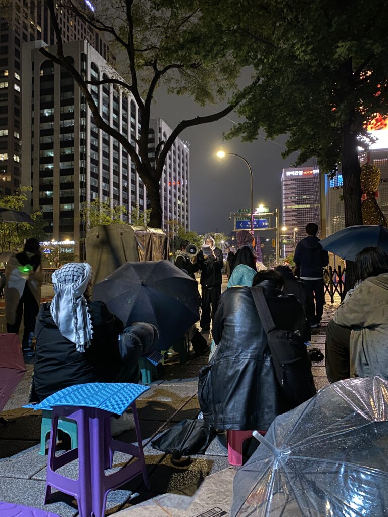 사진2. 오픈마이크 모습. QK48 활동가 화가 팔레스타인 시인의 시를 낭독하고 있다. 옆에서는 확성기를 들어주고 있다.