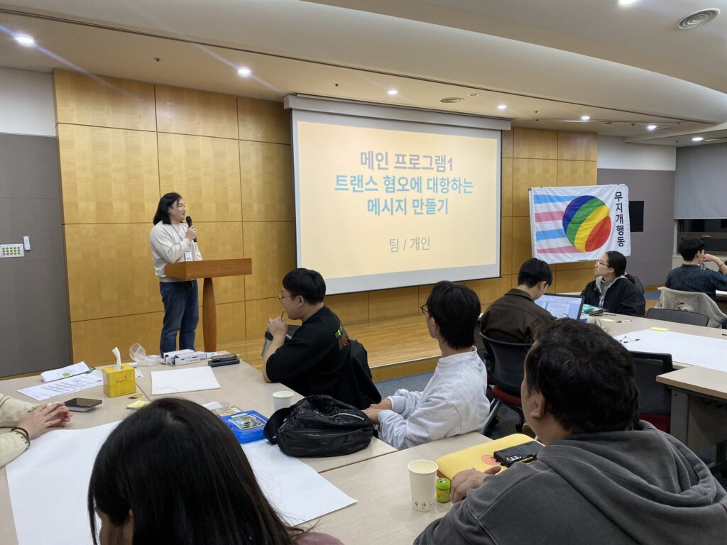 사진1. 소하가 단상 위에서 마이크를 잡고 말하고있다. 스크린에는 메인 프로그램1 트랜스 혐오에 대항하는 메시지 만들기라고 띄워져 있다.