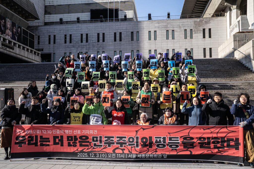 12.10 민중의 행진 선포 기자회견 현장 이미지. 세종문화회관 앞 계단에서 여러 시민·활동가들이 피켓을 들고 모여 있다. 전면에는 ‘우리는 더 많은 민주주의와 평등을 원한다’는 붉은 현수막이 펼쳐져 있다.