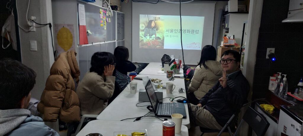 사진1. 서울인권영화클럽 소모임원들이 테이블에 둘러 앉아 스크린을 바라보고 있다. 오른편에 앉은 우진은 카메라를 향해 브이 포즈를 취하고 있다.