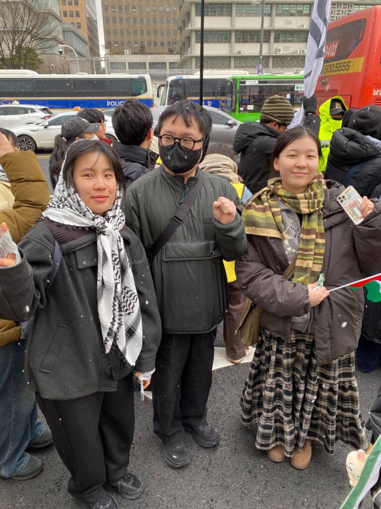 사진. 행진 중인 이음활동가 현토, 우진, 효민이 카메라를 보고 포즈를 취한다. 효민은 작은 팔레스타인 국기를 들고 있다. 함박눈이 내리고 있다.