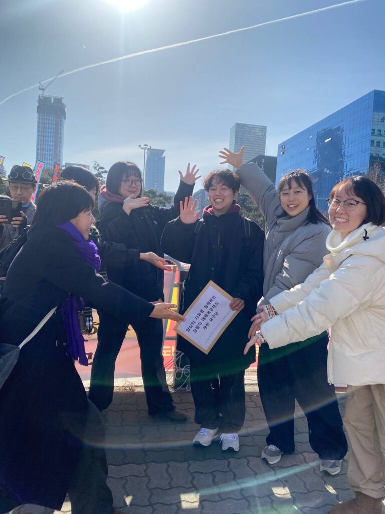 사진1. 송이, 마주, 안나, 고운, 미나상이 두부를 둘러싸고 손바닥으로 두부를 가리키고 있다. 두부는 "양심의 자유를 침해하는 징벌적 대체복무제도 개선 요구안"이 적힌 서류봉투를 들고있다.