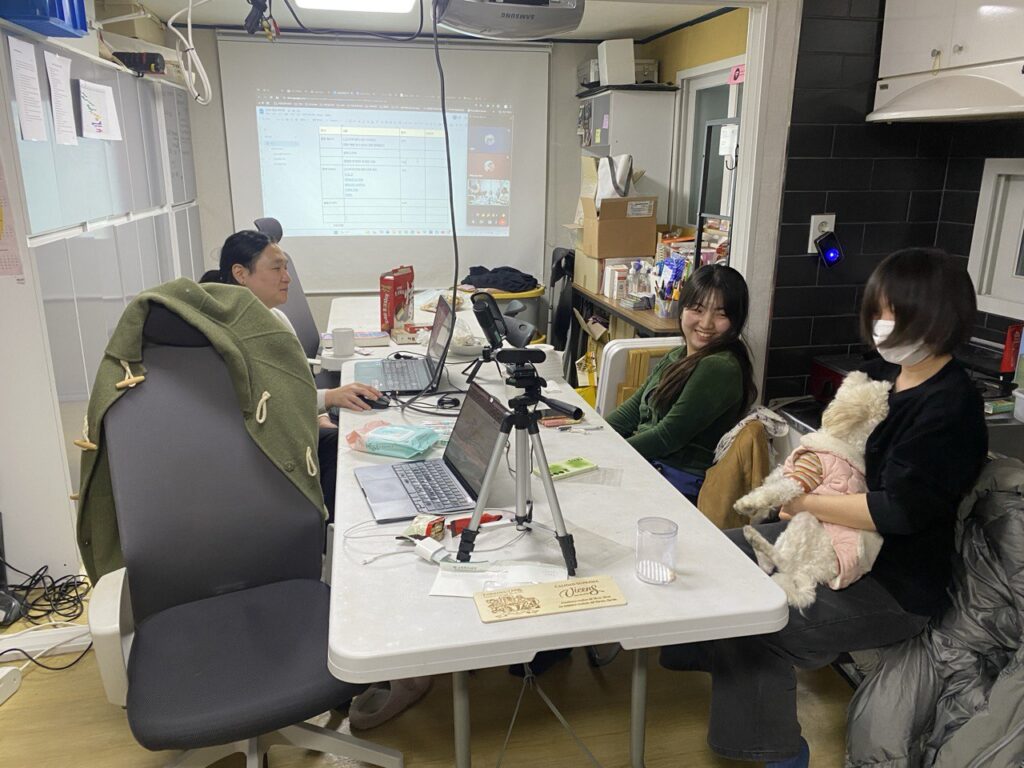 사진1. 테이블 왼쪽 편에는 소하가 앉아있다. 소하는 노트북으로 회의록을 보고있다. 테이블 오른쪽 편에는 지은과 마주가 앉아있다. 마주는 구름이(강아지)를 안고 있고 지은이 구름이를 바라보고 있다.