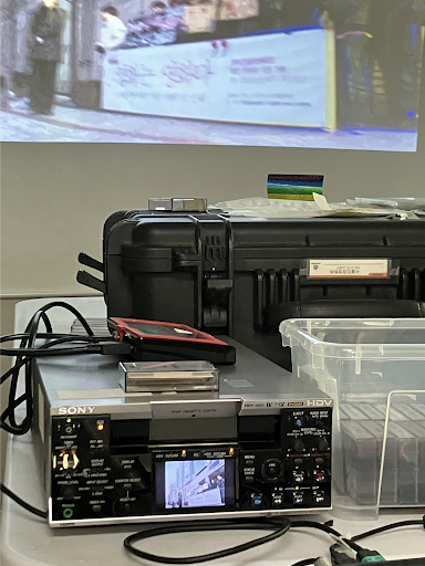 사진 1(왼). 비디오테이프 플레이어로 서울인권영화제 옛 활동 기록을 살펴보고 있는 모습. 왼편에는 비디오테이프 플레이어가, 오른편에는 비디오테이프 더미가 담긴 박스가, 그 뒤에는 스크린이 있다.