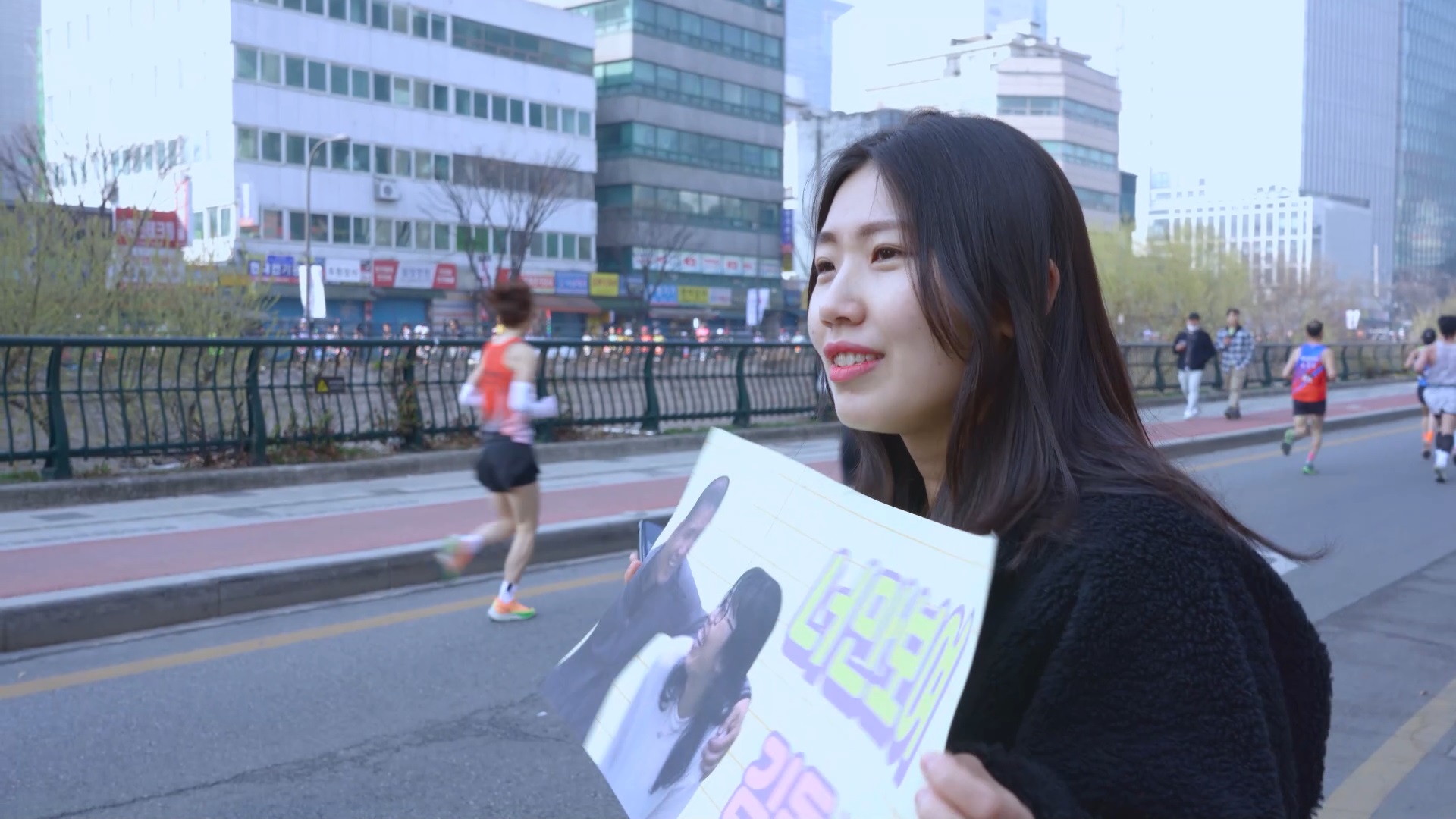 스틸컷6. 한 사람(김동수의 딸)이 플랜카드를 펼쳐들고 서있다. 플랜카드엔 두 사람이 웃으며 어깨동무를 하는 사진과 너만보여 … 까지 문구가 적혀있다. 뒤로는 운동복 차림을 한 마라토너들이 도로를 달리고 있다.