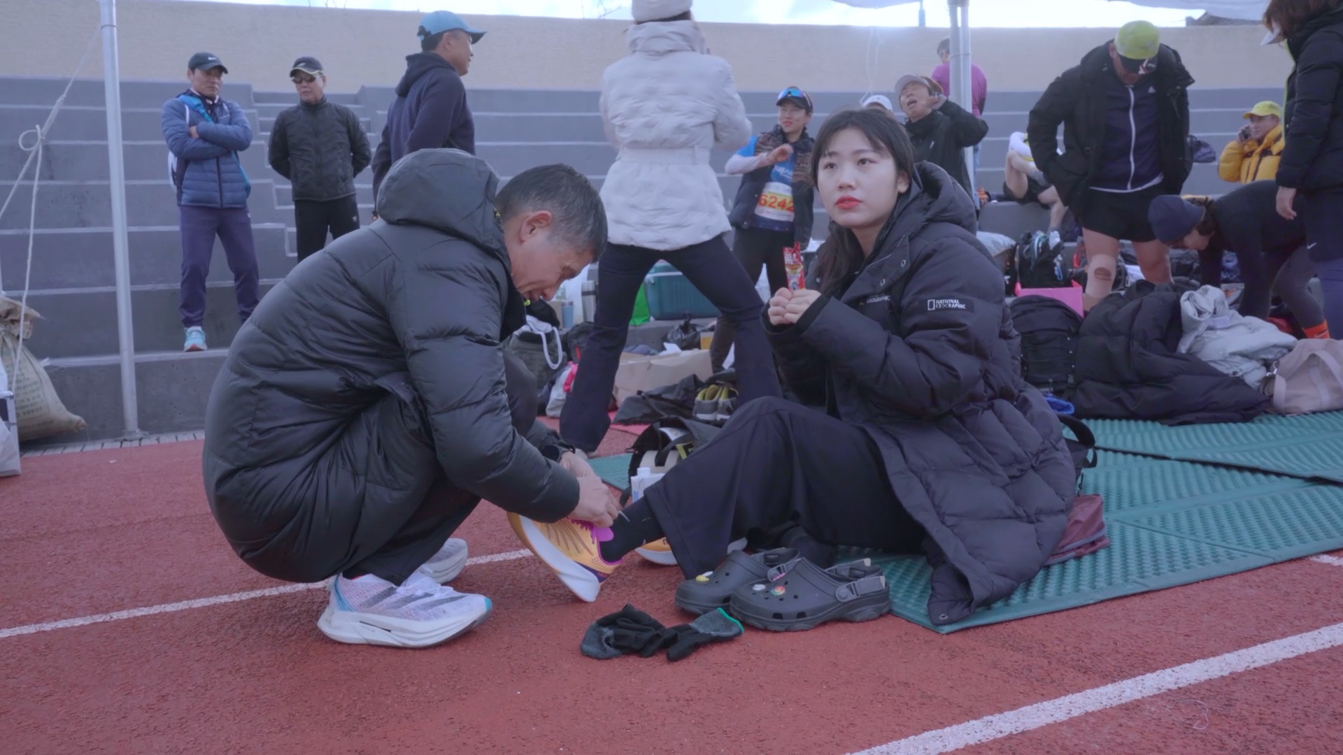 스틸컷8. 두 손으로 에너지젤을 들고 바닥에 앉아 발을 뻗고 있는 딸. 그 맞은 편에 있는 짧은 머리의 중년 김동수가 쪼그려 앉아 뻗어진 쪽의 신발끈을 묶고 있다. 뒤로는 운동복 차림의 사람들이 서서 몸을 풀거나 복장을 정돈하고 있다.