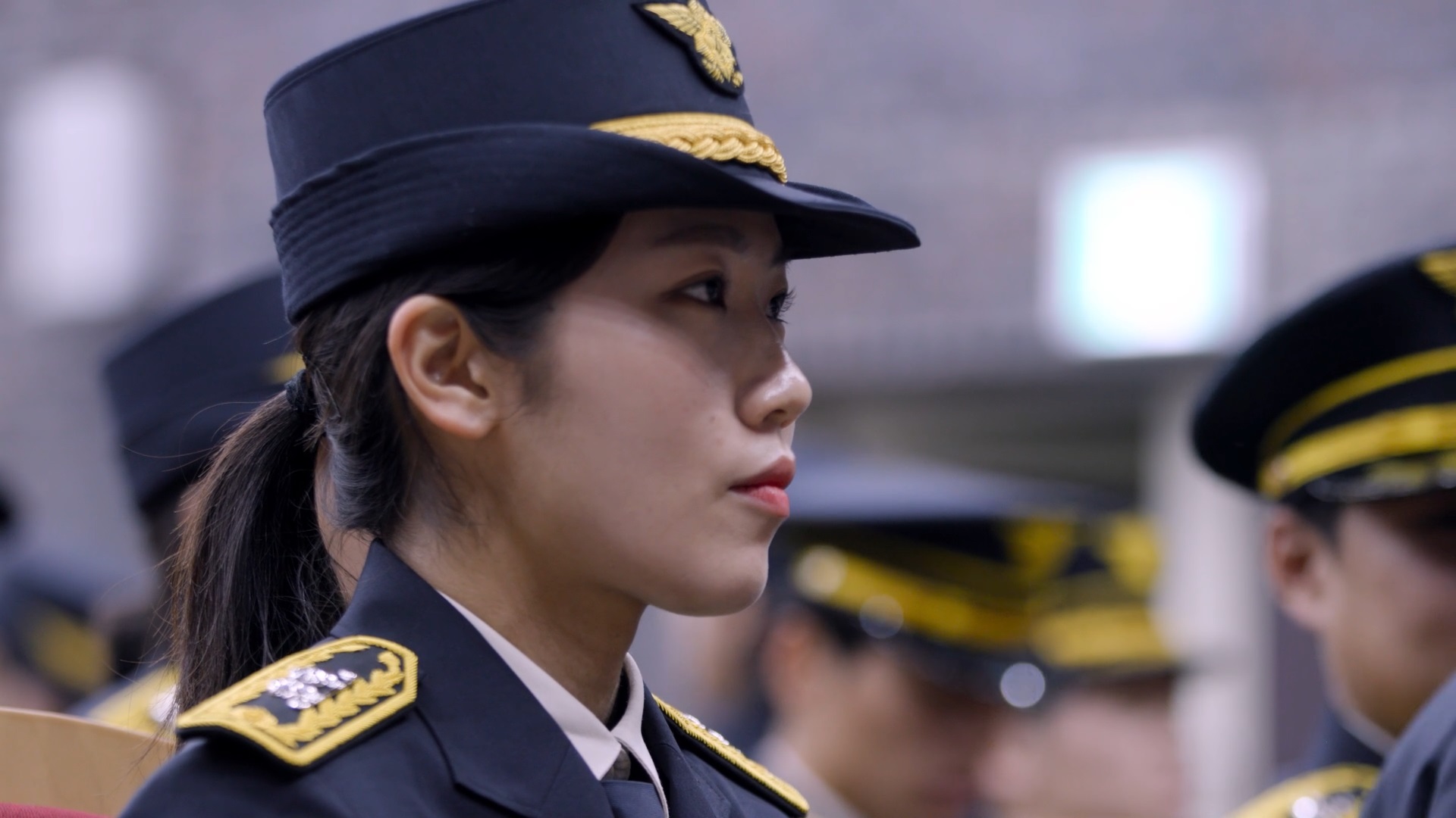 스틸컷9. 소방정복을 입은 사람의 옆모습. 단정하게 머리를 묶고 결연한 표정이다. 뒤로는 똑같은 정복을 입은 사람들이 있다.