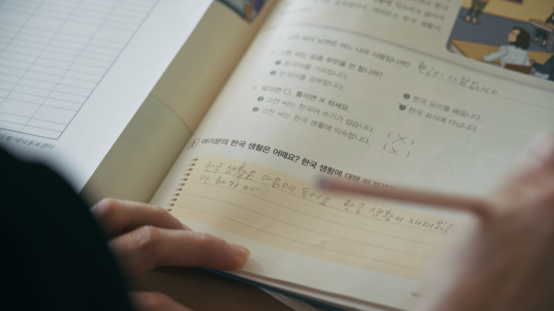 스틸컷3. 문제를 풀고 있는 연필을 쥔 손이 있다. 문제지에는 ‘여러분의 한국 생활은 어때요?’라고 적혀있다. 답변을 쓰는 칸에는 필기체로 한국 생활은 마음에 들어요 한국 생활이 재미있•••말하기 어•• 까지 적혀있다.