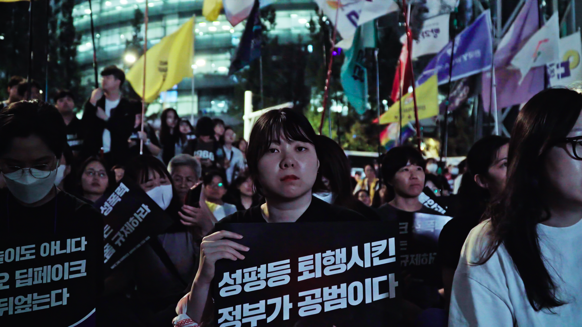 스틸컷1. ‘성평등 퇴행시킨 정부가 공범이다’ 라는 피켓을 들고 있는 한 사람이 카메라를 응시하고 있다. 그 주변에는 많은 사람들이 시위에 참여하고 있다. 여러 연대체의 깃발들이 있다.