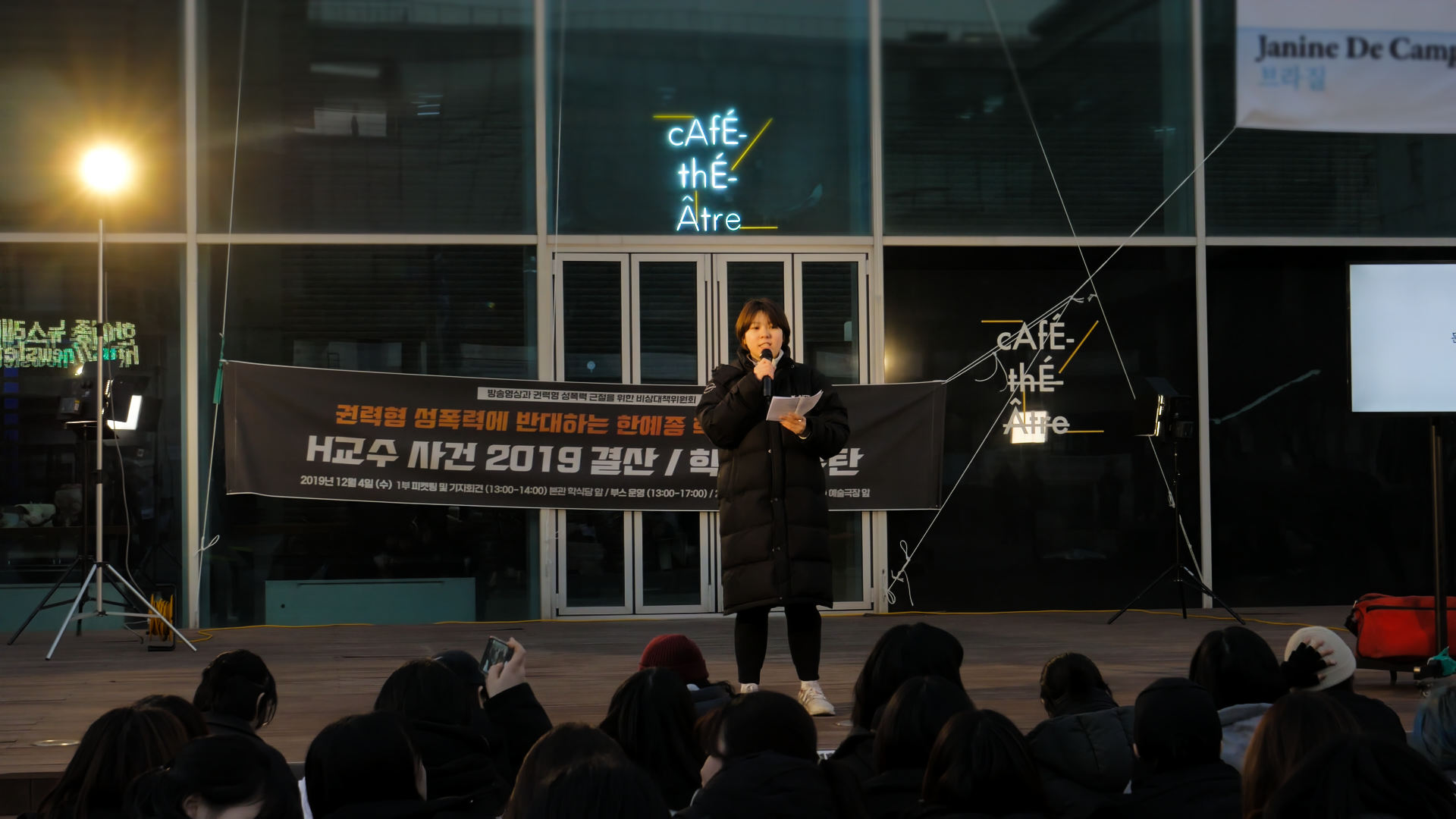 스틸컷2. ‘권력형 성폭력에 반대하는 한예종-’ 현수막이 달려있고 그 앞에 롱패딩을 입은 한 사람이 단상 위에서 연설을 하고 있다. 단상아래 연설자를 바라보고 있는 사람들의 뒤통수.