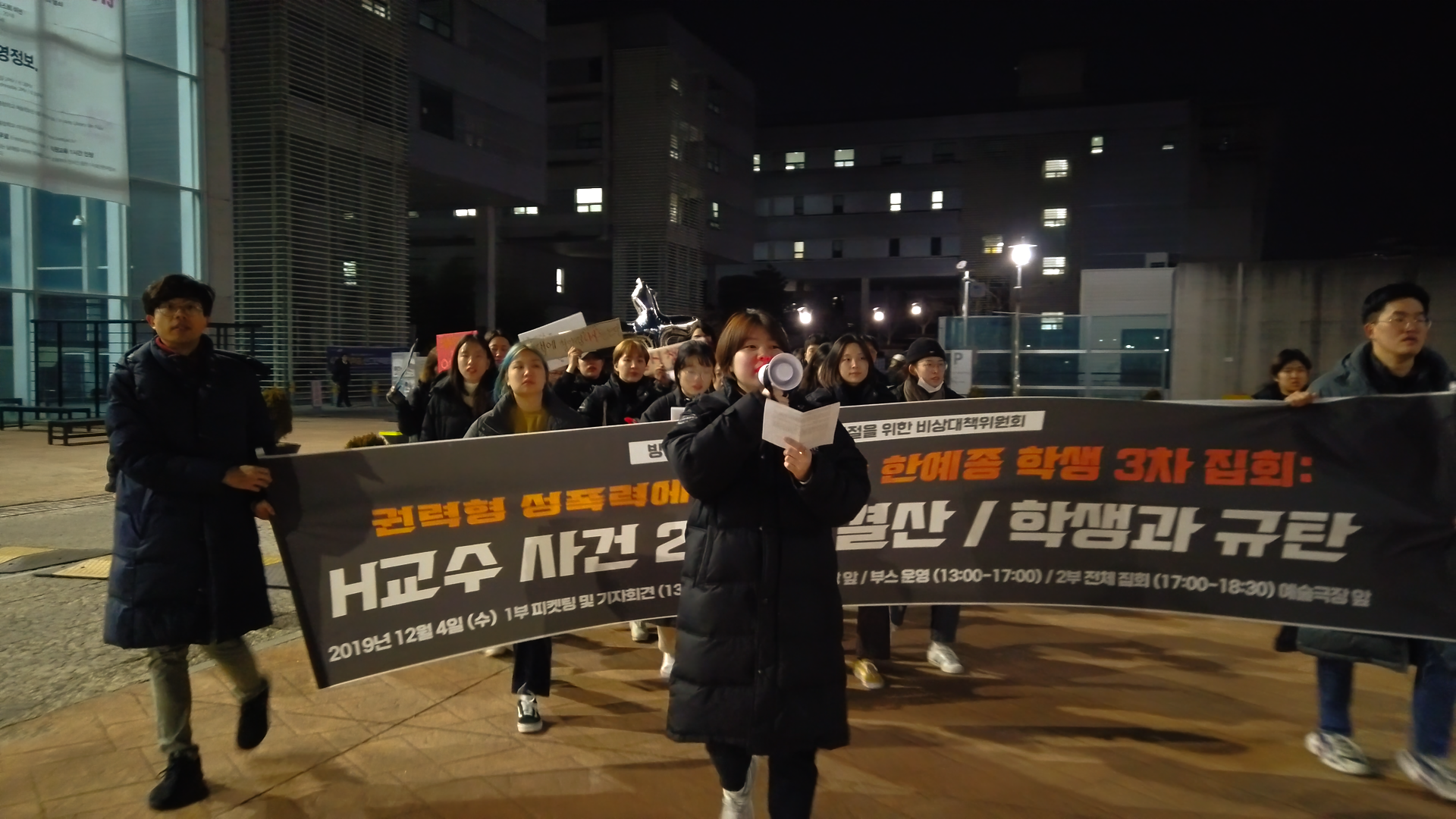 스틸컷7. ‘권력형 성폭력, 한예종 학생 3차 집회, 학생과 규탄’이 적혀있는 현수막을 양쪽에서 두 명이 잡고 걸어오고 있다. 그 현수막 앞에서 선두로 확성기를 든채 말하며 걸어오는 사람 한 명이 있다. 현수막 뒤를 따라오는 행진하고 있는 사람들이 있다.