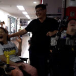 스틸컷1. 전동 휠체어를 탄 두 사람 가운데 검은 티셔츠를 입고 안경을 쓴 사람이 있다. 셋이 환하게 웃고 있다. 왼쪽 사람은 흰색 반팔티와 분홍 긴바지를 입고 있다. 오른쪽 사람은 검은 반팔티를 입고 있다.