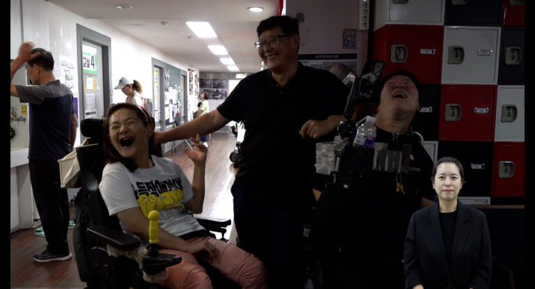 스틸컷1. 전동 휠체어를 탄 두 사람 가운데 검은 티셔츠를 입고 안경을 쓴 사람이 있다. 셋이 환하게 웃고 있다. 왼쪽 사람은 흰색 반팔티와 분홍 긴바지를 입고 있다. 오른쪽 사람은 검은 반팔티를 입고 있다.