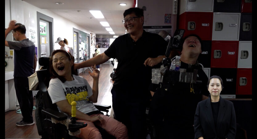 스틸컷1. 전동 휠체어를 탄 두 사람 가운데 검은 티셔츠를 입고 안경을 쓴 사람이 있다. 셋이 환하게 웃고 있다. 왼쪽 사람은 흰색 반팔티와 분홍 긴바지를 입고 있다. 오른쪽 사람은 검은 반팔티를 입고 있다.