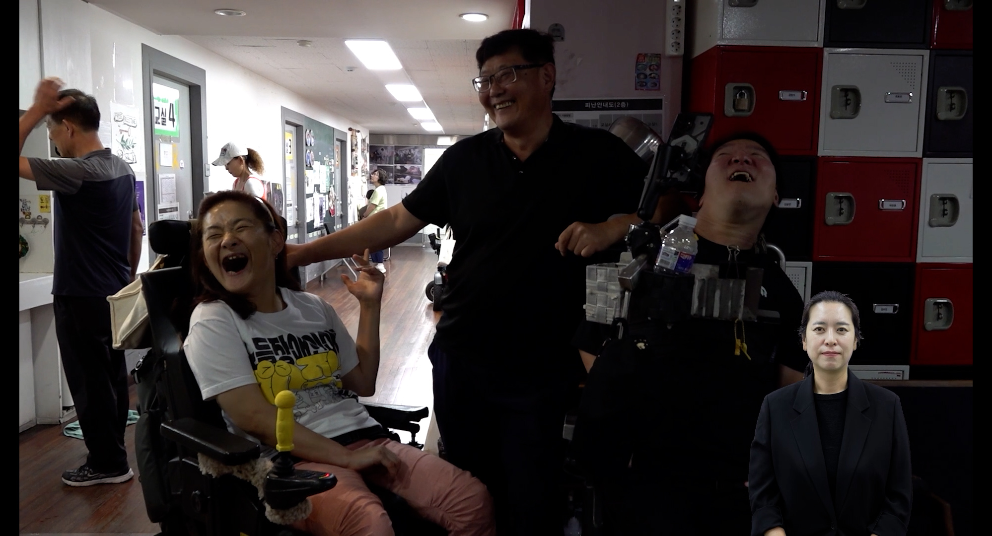 스틸컷1. 전동 휠체어를 탄 두 사람 가운데 검은 티셔츠를 입고 안경을 쓴 사람이 있다. 셋이 환하게 웃고 있다. 왼쪽 사람은 흰색 반팔티와 분홍 긴바지를 입고 있다. 오른쪽 사람은 검은 반팔티를 입고 있다.