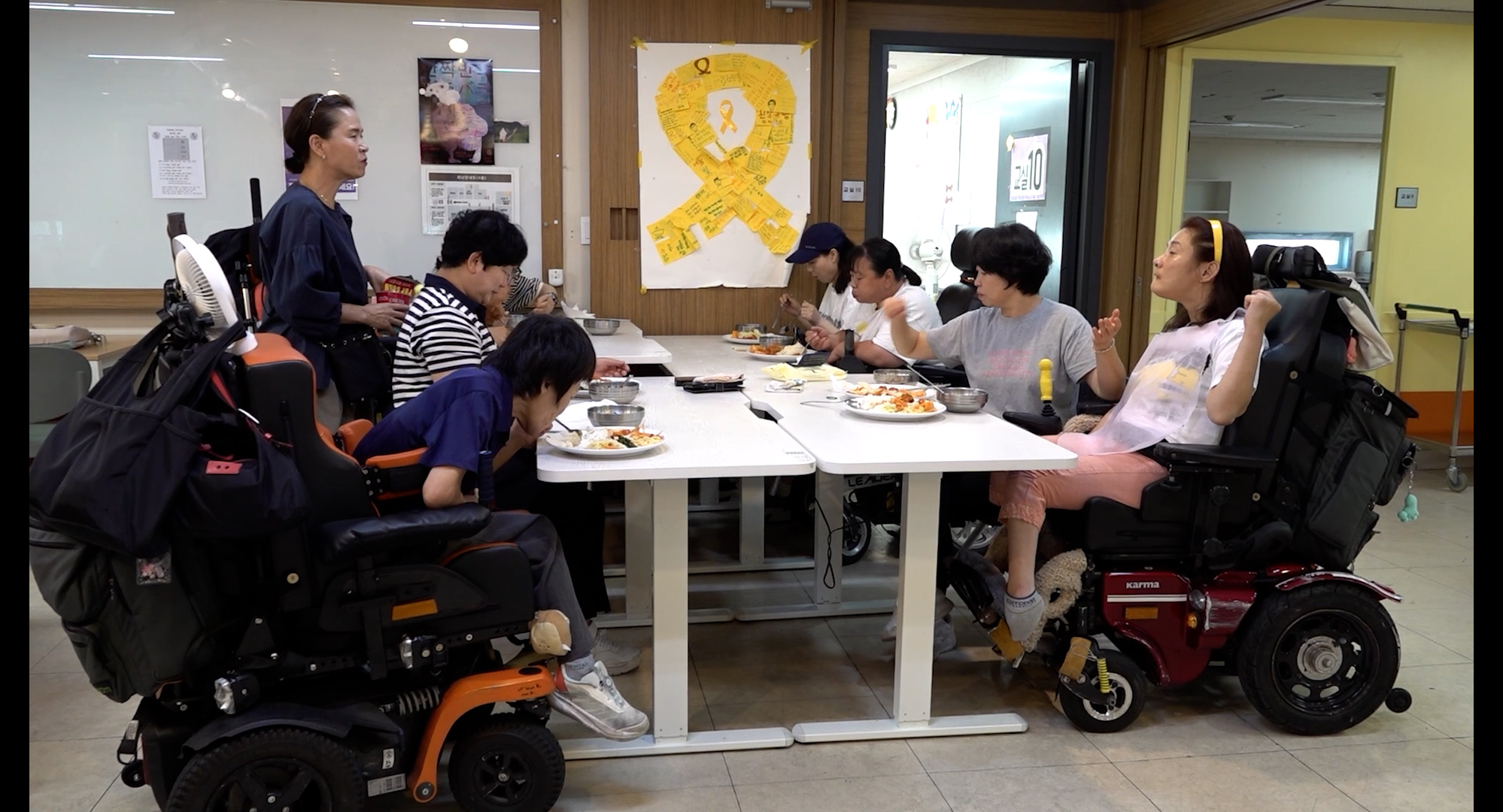 스틸컷3. 8명의 사람이 4명씩 마주보며 흰 책상 위에서 밥을 먹고 있다. 한 사람은 일어나서 그들과 대화하고 있다. 그들 뒤로 보이는 벽에는 글씨가 적혀있는 노란 메모지 여러개가 큰 노란 리본 형상으로 붙여져있다.