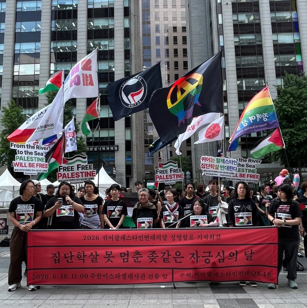 사진1. 기자회견 현장. ‘2026 퀴어팔레스타인연대의달 성명발표 기자회견 집단학살 못 멈춘 좆같은 자긍심의 달’이 적힌 붉은 현수막 뒤로 참여자들과 참여 단체의 깃발들이 서 있다. 참여자들은 검은 옷을 입었다. 티셔츠에는 팔레스타인 국기와 프라이드플래그가 인쇄된 스티커를 붙였다.