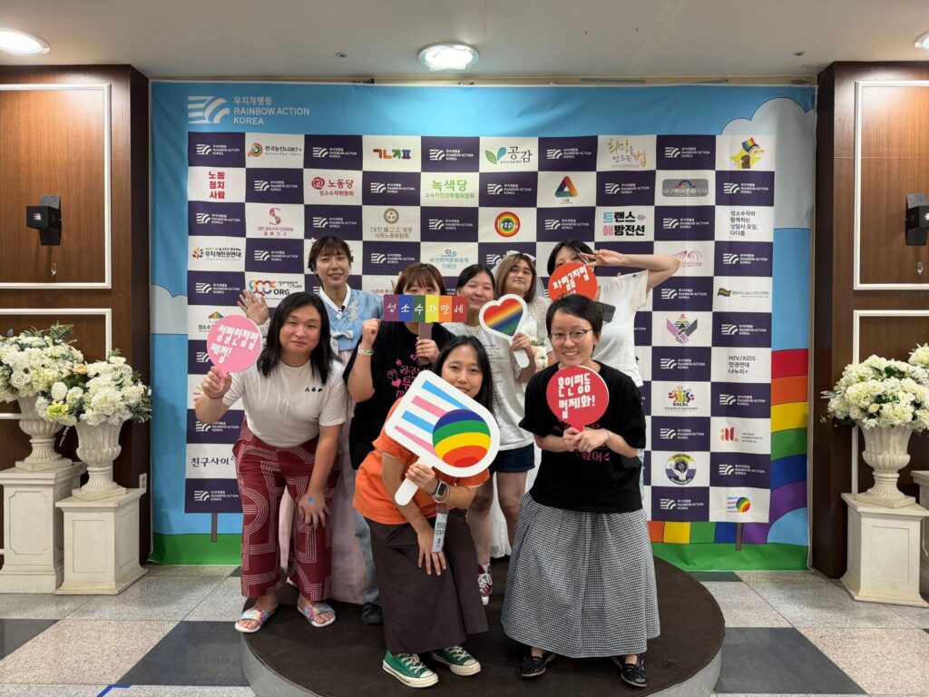 사진1. 연대의밤 행사장 입구에 마련된 포토월에서 서울인권영화제 활동가들의 단체 사진. 맨 뒤에 트랜스 플래그 색상의 혼주 한복을 입은 박한희 무지개행동 공동대표. 활동가들은 '성별인정법 제정', '차별금지법 제정', '혼인평등 법제화' 등 피켓을 들고 있다. 배경 현수막에 무지개행동 소속단체들의 로고가 인쇄되어있다.