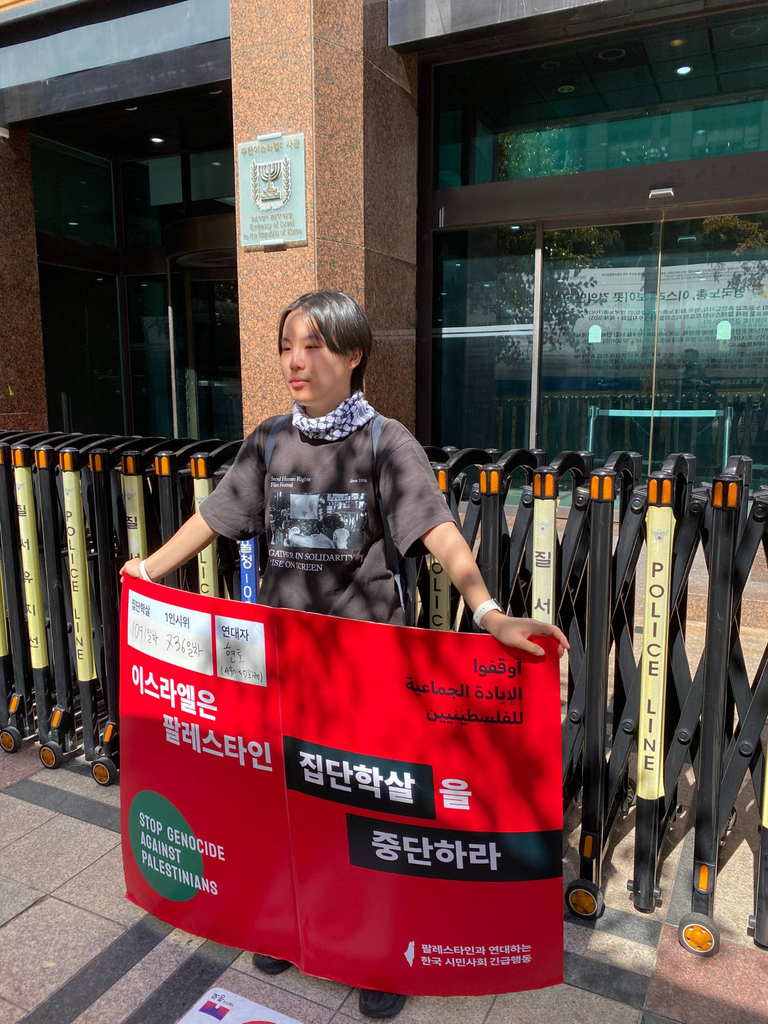 이스라엘 대사관 앞에서 1인 시위를 하고 있는 현토. 직사각형 모양의 빨간색 피켓을 들고 서 있다. 피켓 상단에는 연대자 현토의 이름과 함께 집단학살 1091일차, 1인 시위 736차라는 문구가 써 있다. 또, 피켓 하단에는 “이스라엘은 팔레스타인 집단학살을 중단하라”는 문구가 써 있다.