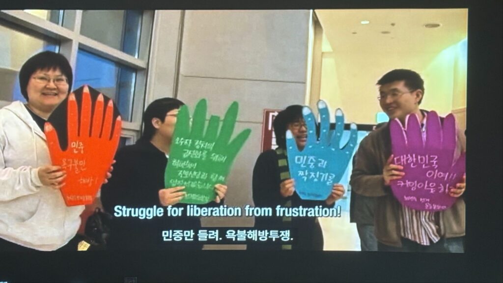[사진]. 영화제 사무실 스크린 속 ⟨레즈비언정치도전기⟩의 한 장면. 선거 운동하는 사람 네 명이 손 모양의 팻말을 들고 있다. 팻말에는 “민중 욕구불만해방!”, “소수자 정치의 급진화를 위해 진보신당과 함께 앞장 서겠습니다!”, “민중과 짝짓기를”, “대한민국이여!! 커밍아웃하라!!” 가 적혀있다.
